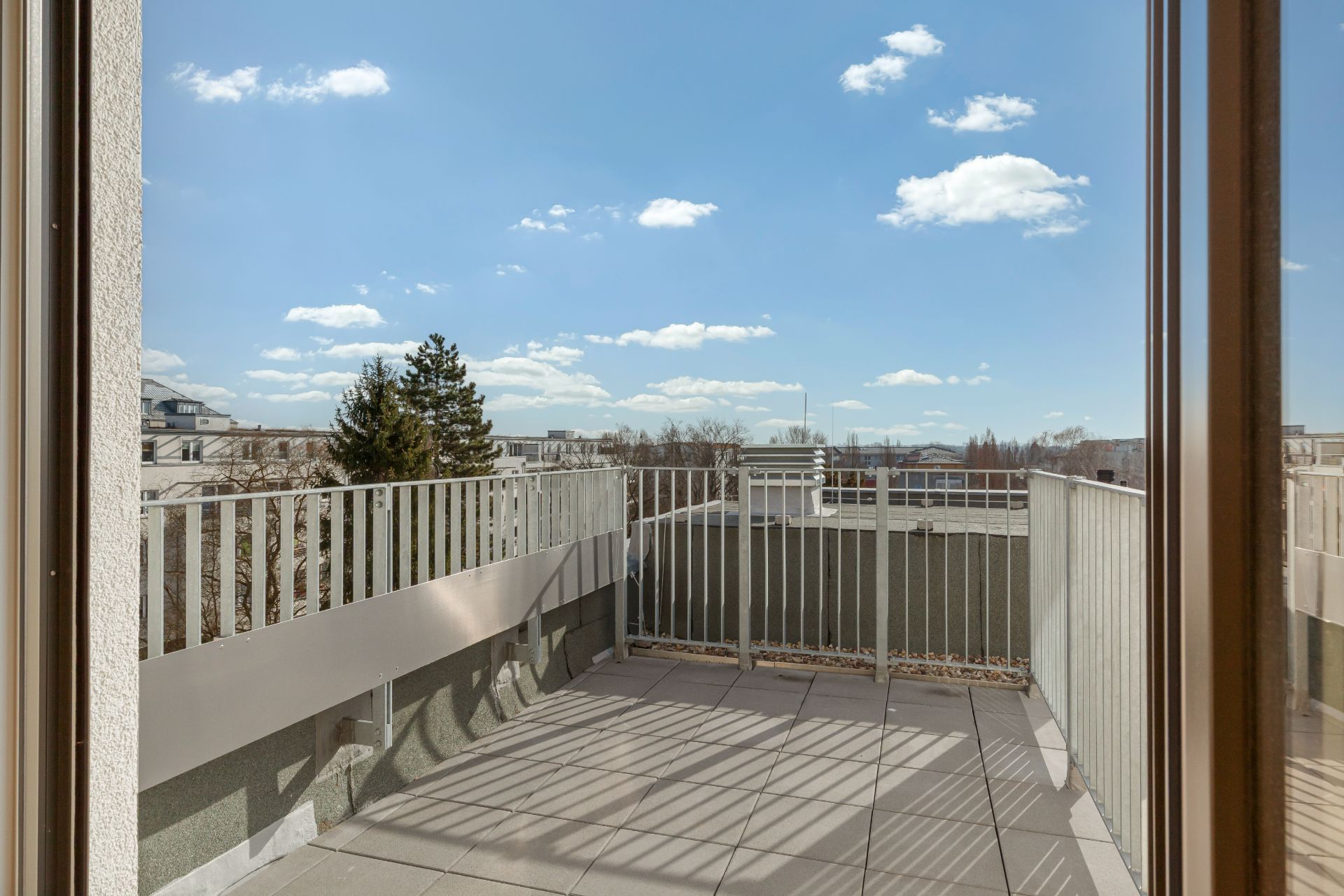 Blick von innen auf einen Balkon mit Geländer und Fliesenboden, im Hintergrund blauer Himmel.