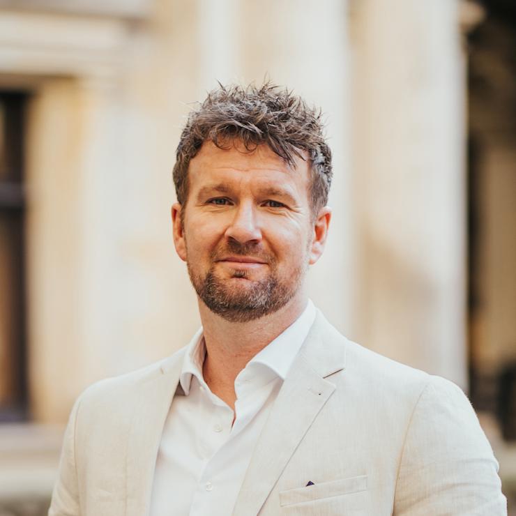 Ein Mann mit hellbeigem Sakko und weißem Hemd steht draußen vor unscharfem Hintergrund.