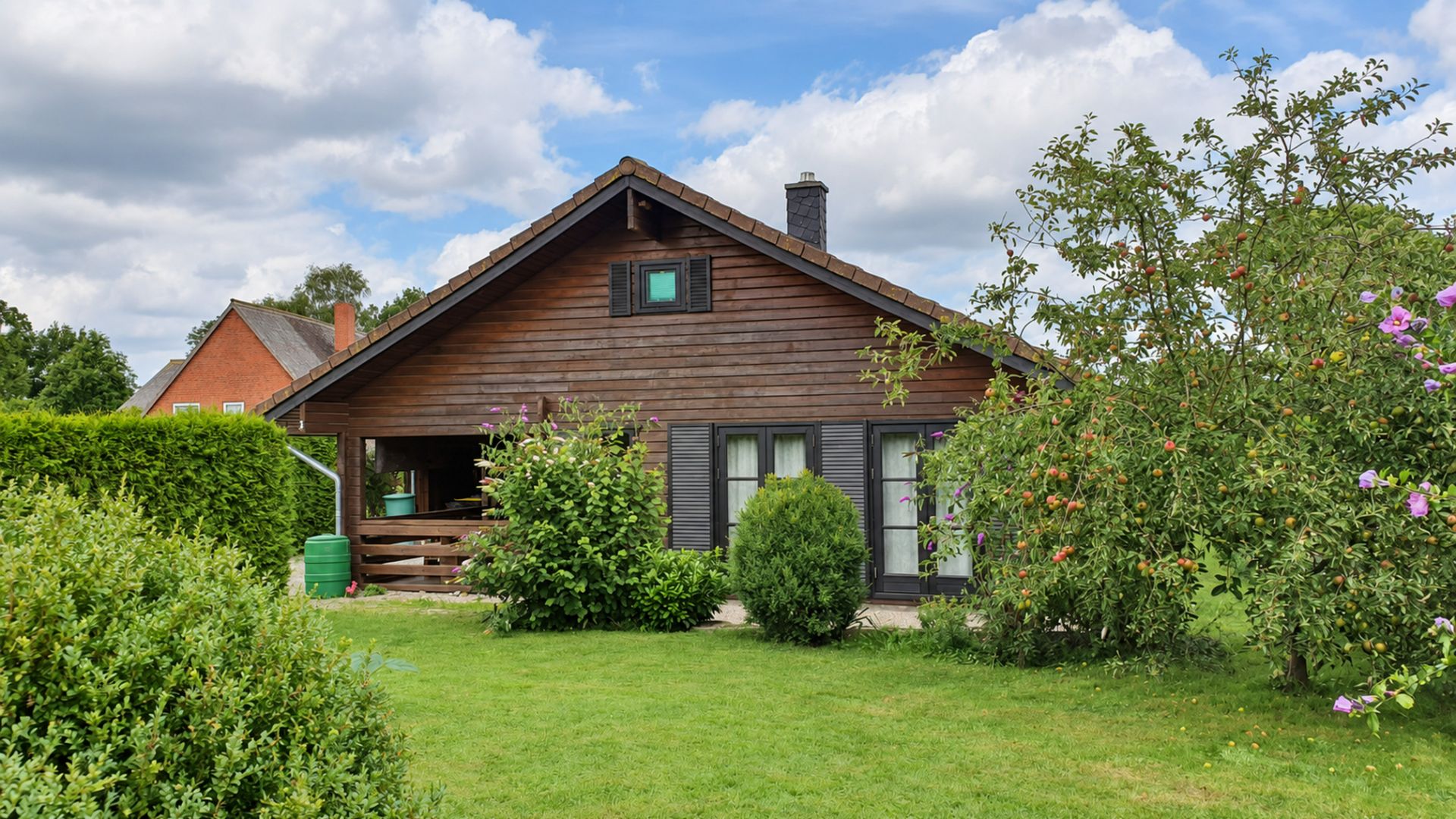 Holzhaus mit Garten, Sträuchern und Bäumen, davor Rasenfläche.