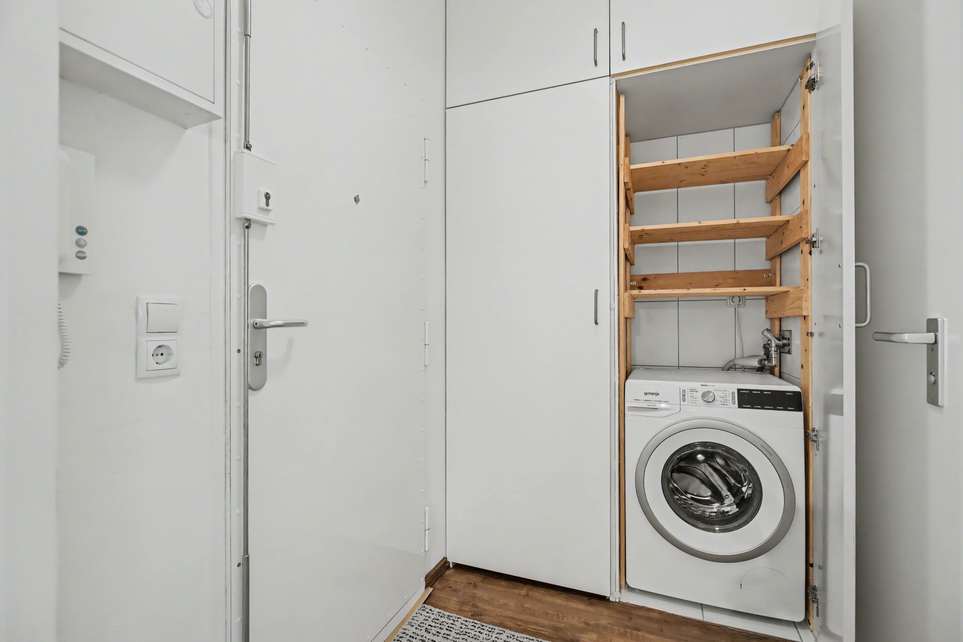 Waschmaschine steht in einem offenen weißen Einbauschrank mit Holzregalen in einer hellen Ecke.