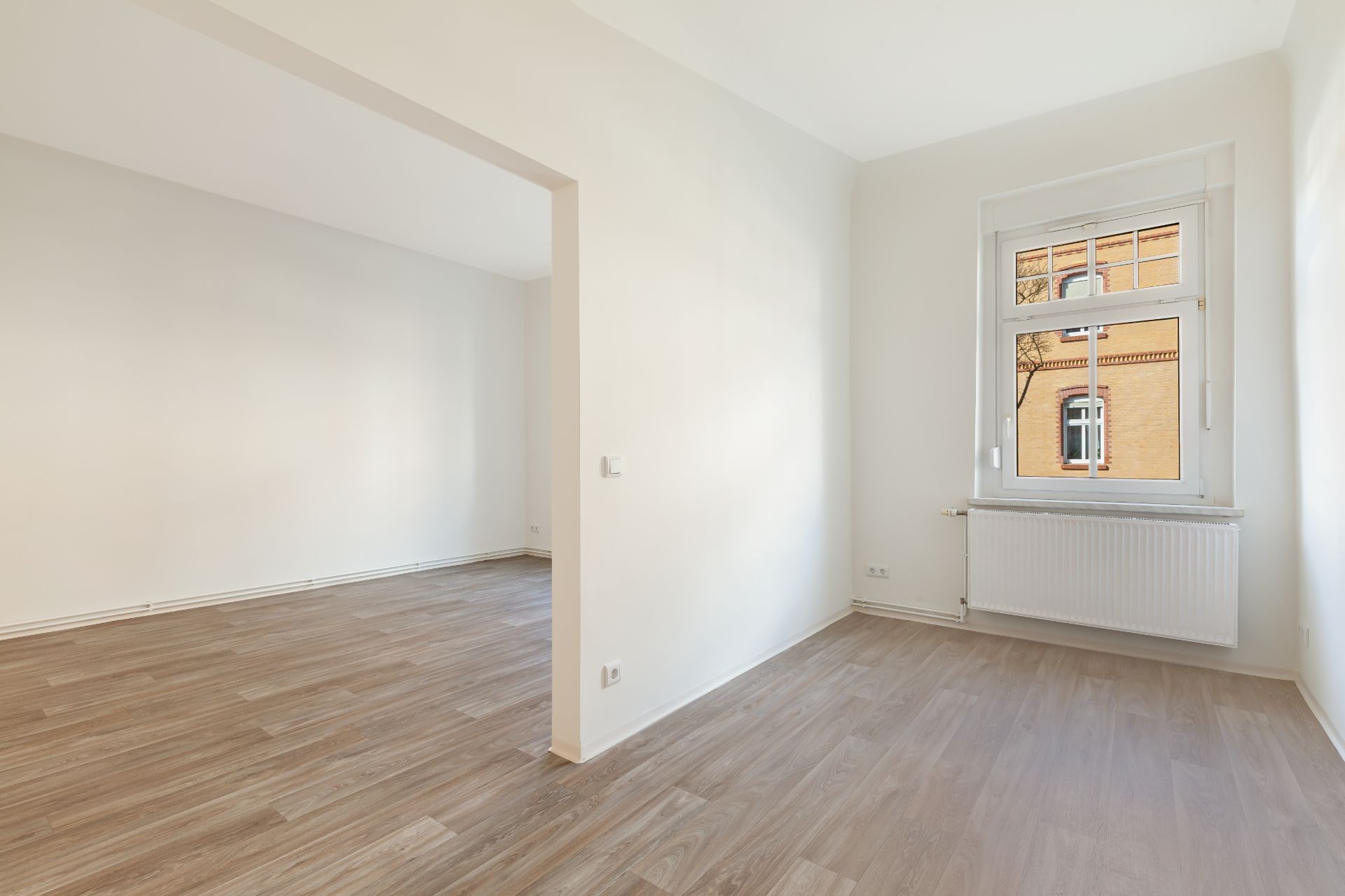 Heller Raum mit großem Fenster, Heizkörper und Holzfußboden, Wandöffnung zu weiterem Zimmer.