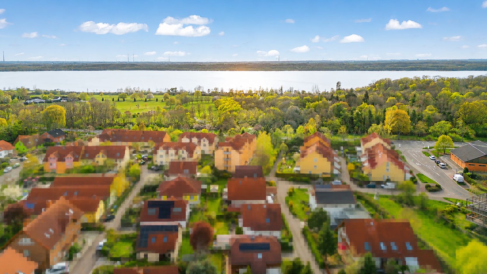 Luftaufnahme eines Wohngebiets mit roten Dächern, grünem Park und Fluss im Hintergrund.
