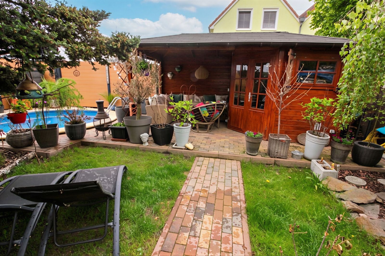Garten mit Rasen, Liegen, vielen Blumentöpfen und einer überdachten Holzterrasse vor einem Haus.
