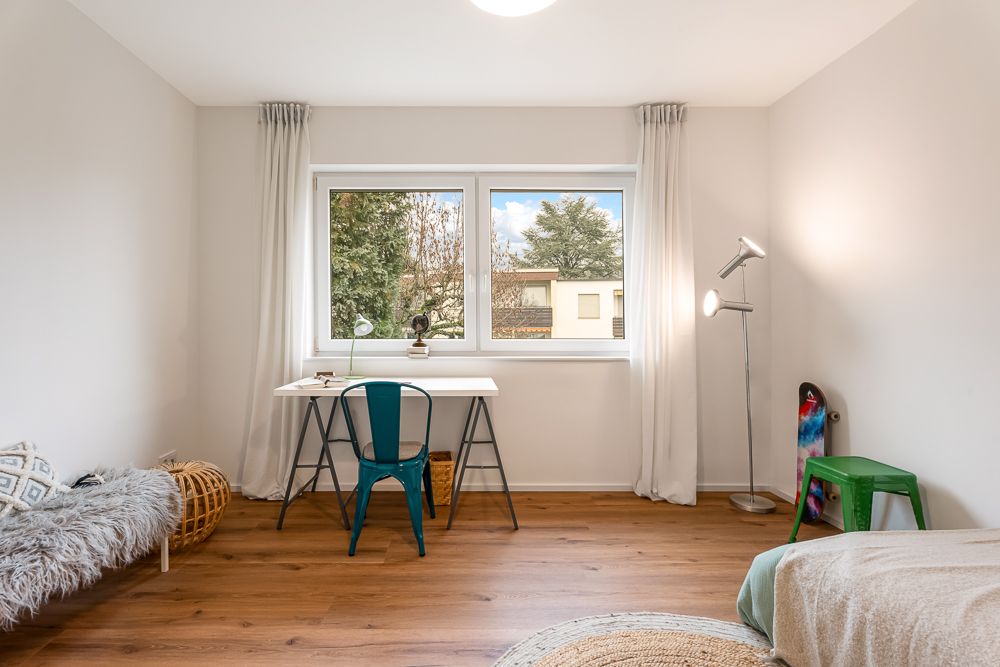 Helles Zimmer mit Schreibtisch am Fenster, Stuhl, Stehlampe, Skateboard und Bett.