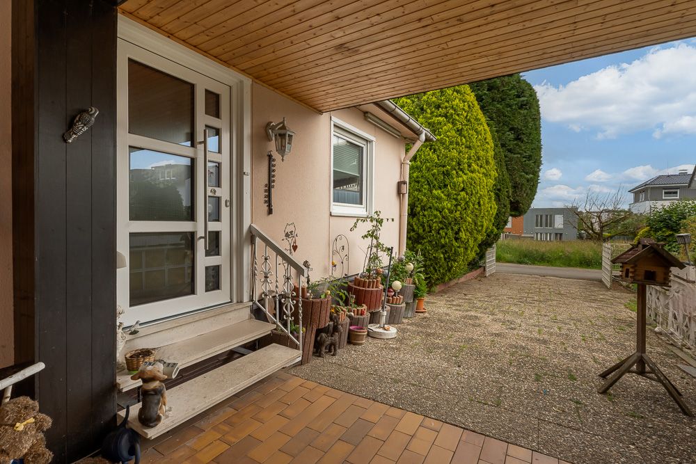 Überdachter Eingang mit Treppe, Pflanzen in Töpfen und gepflastertem Hof vor einem Wohnhaus.