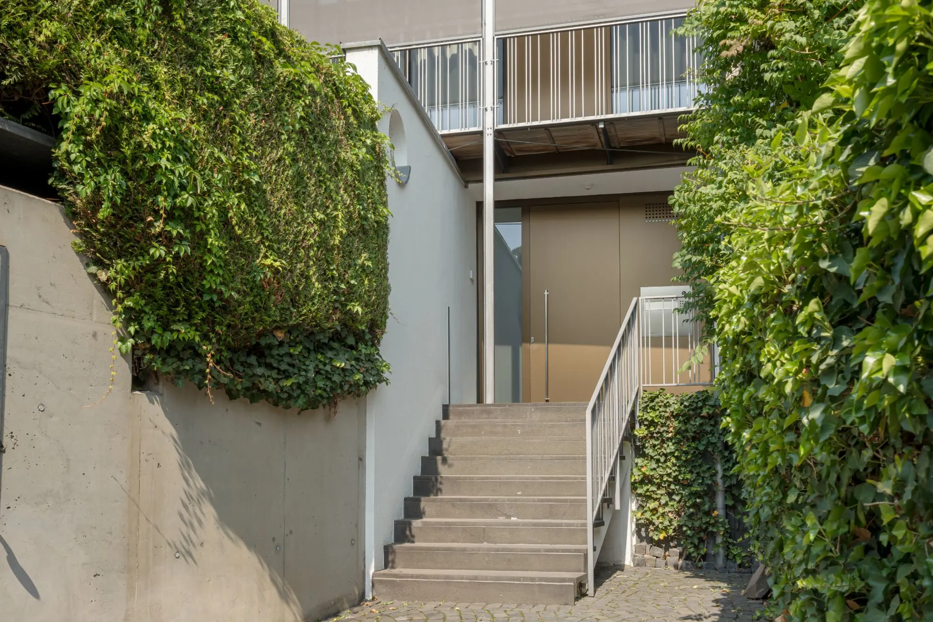Treppe mit Metallgeländer führt zu einer Eingangstür, umgeben von grünen Hecken und Kletterpflanzen.