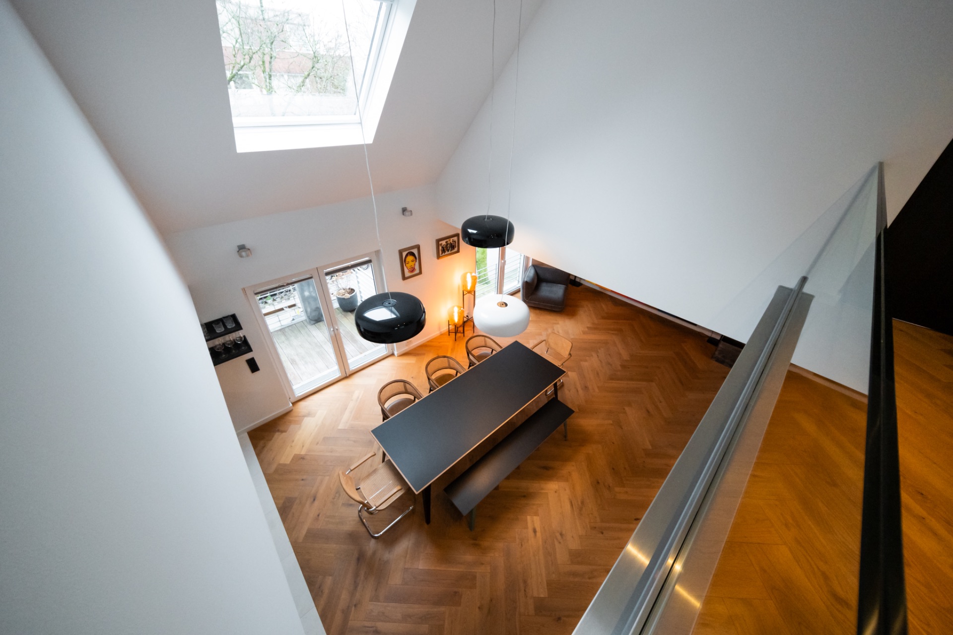 Wohnzimmer mit Esstisch, Stühlen, Bank, Parkettboden, großen Fenstern und Hängelampen.