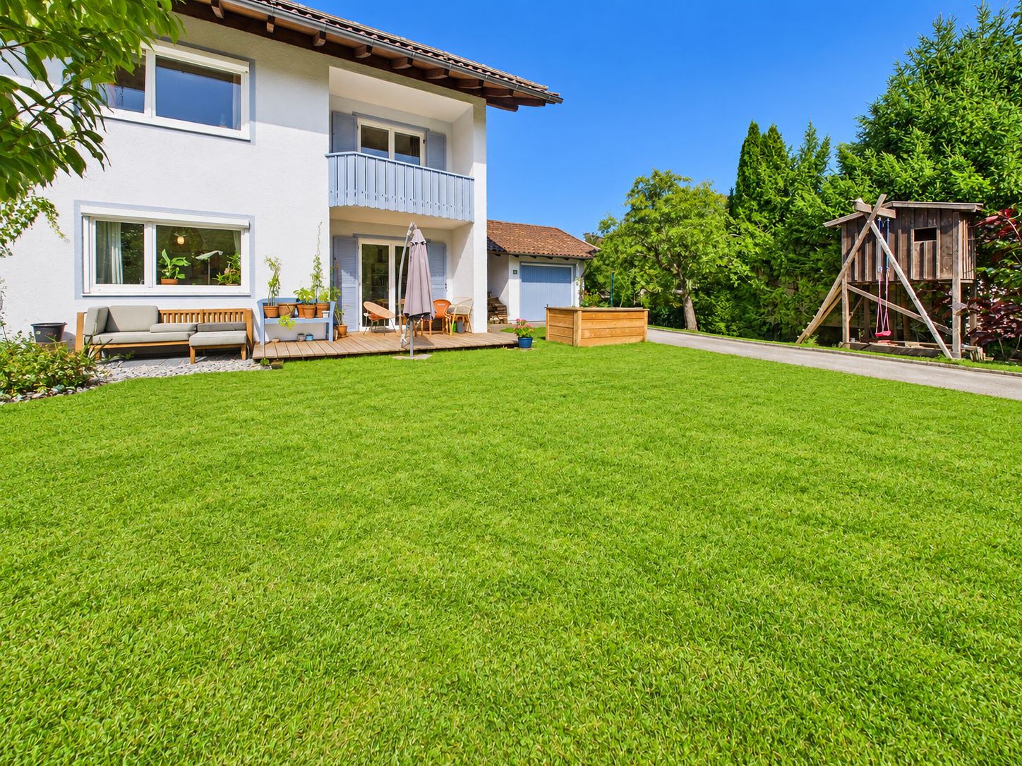Modernes Wohnhaus mit Balkon, Terrasse, großem Garten und Holzspielhaus im Hintergrund.