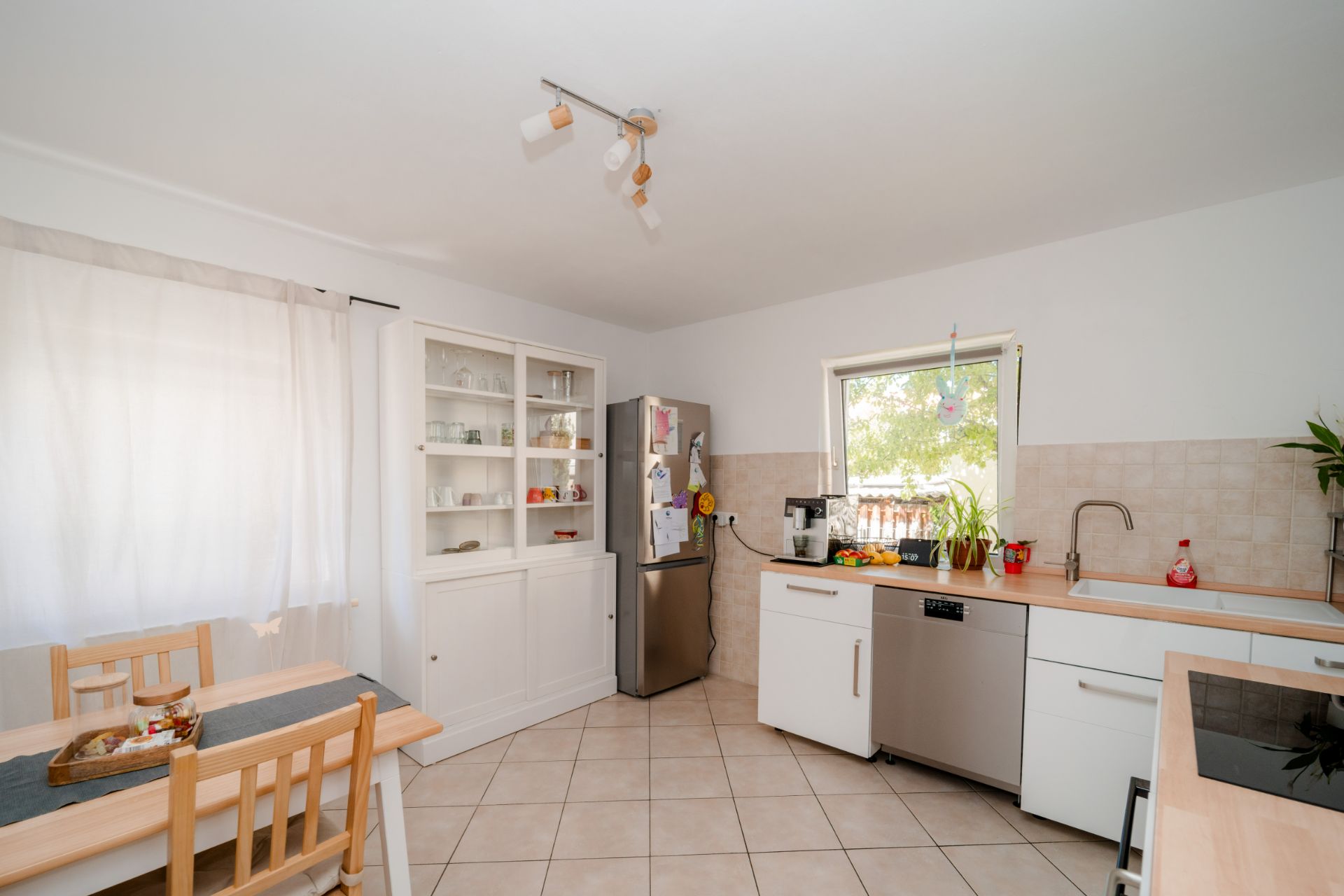 Helle Wohnküche mit Esstisch, weißen Schränken, Kühlschrank und Fenster.