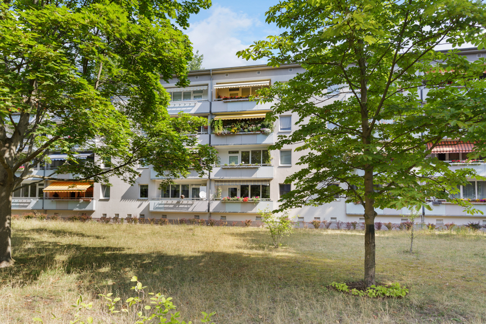 Mehrstöckiges Wohnhaus mit Balkonen und Sonnenschutz, Bäume und Wiese im Vordergrund.