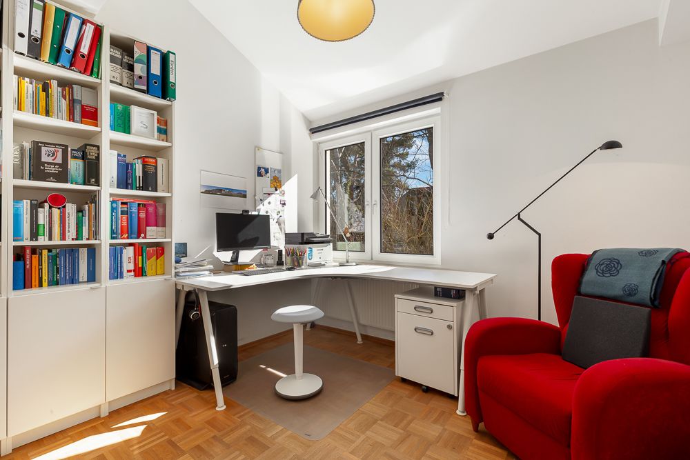 Helles Arbeitszimmer mit Schreibtisch, Bürostuhl, Bücherregal und rotem Sessel am Fenster.