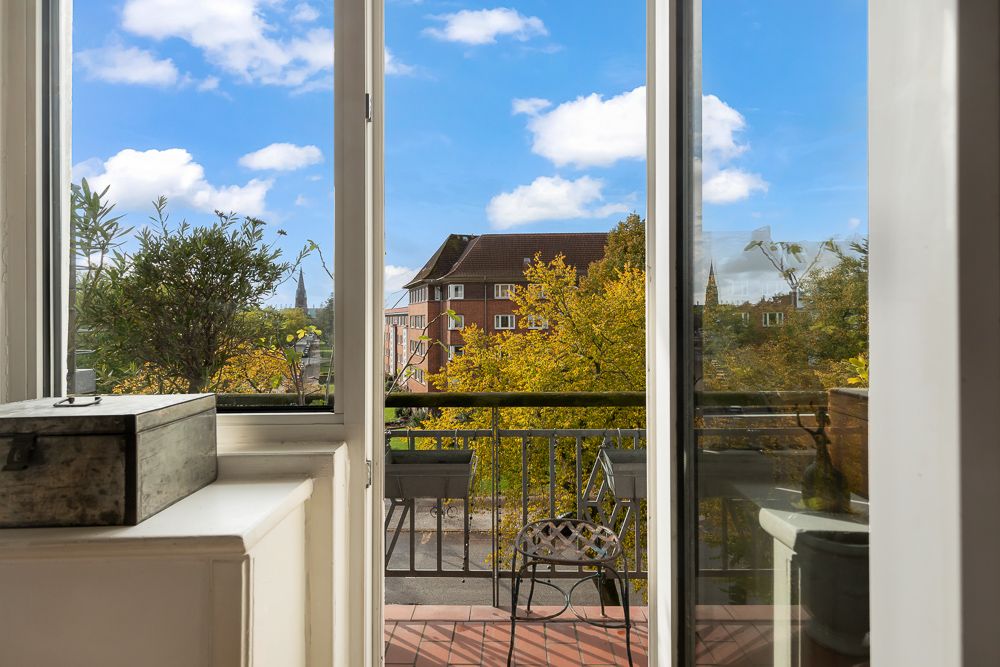 Blick durch geöffnete Balkontür auf Balkon mit Stuhl und Pflanzen, im Hintergrund Bäume und Wohngebäude.