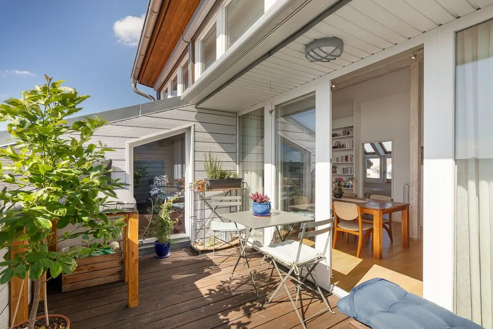Balkon mit Holzboden, Tisch, Stühlen und Pflanzen, Zugang zu einem hellen Esszimmer.