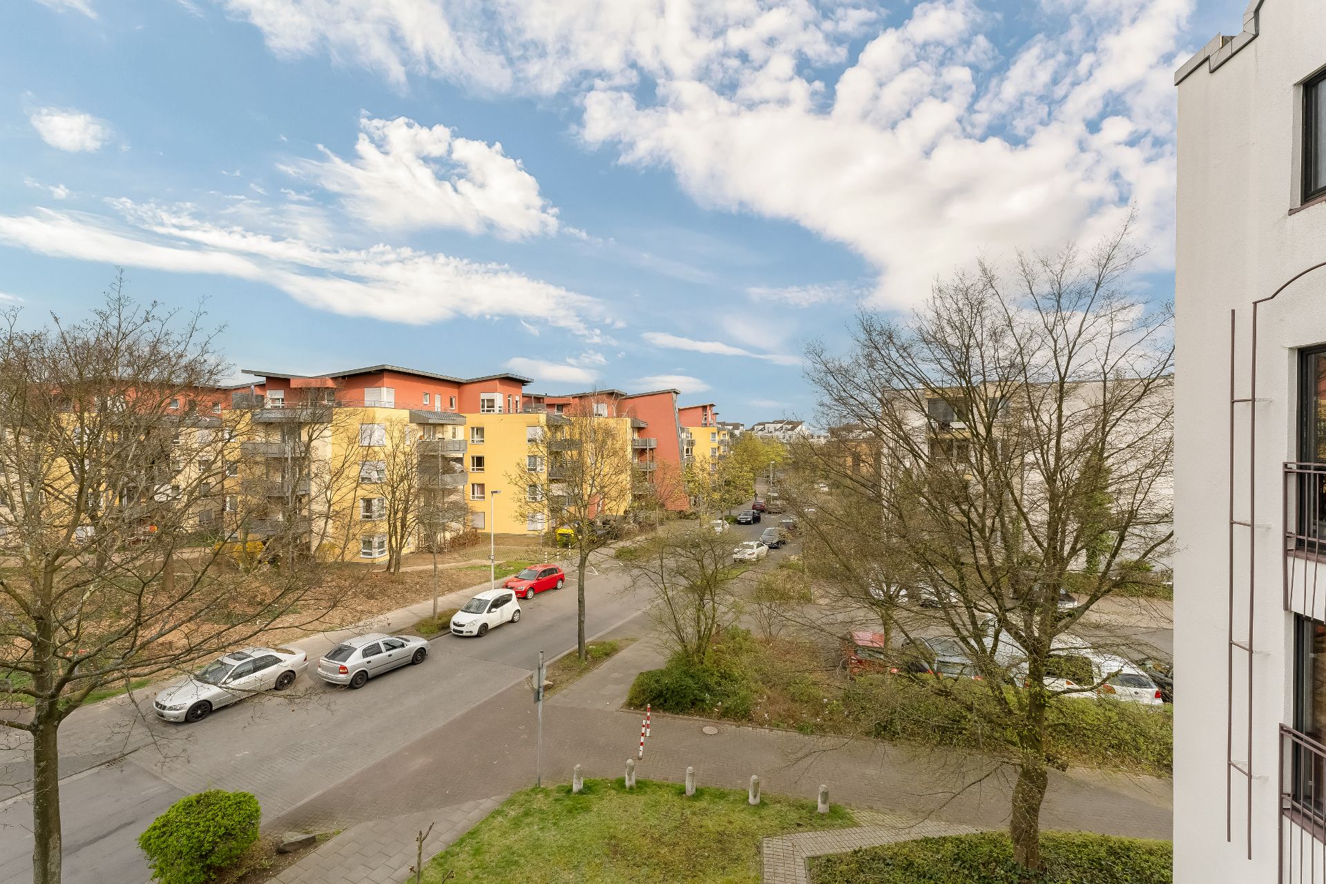 Wohnstraße mit parkenden Autos, Bäumen und mehrstöckigen Wohngebäuden an einem sonnigen Tag.