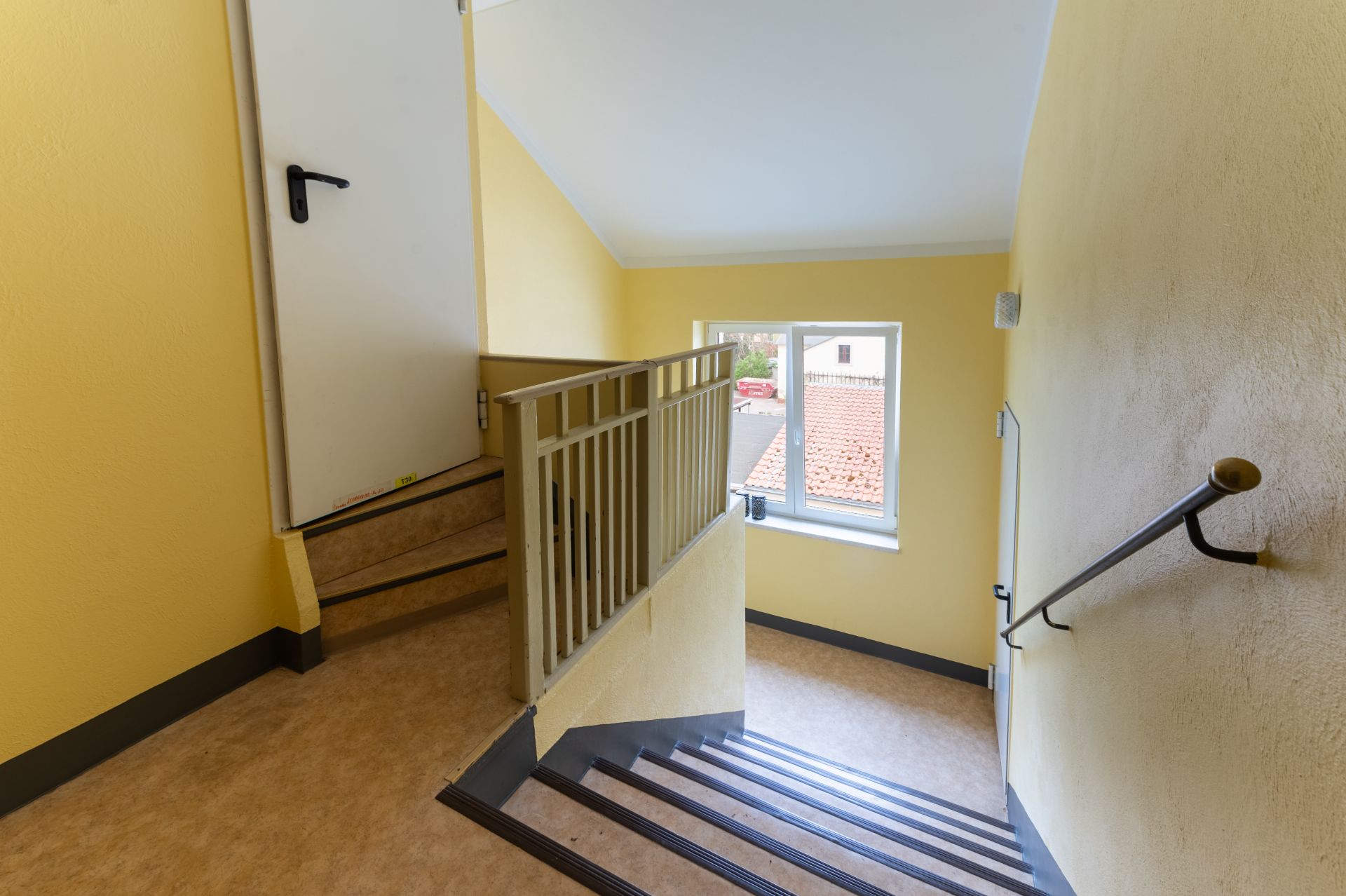 Gelbes Treppenhaus mit Fenster und weißer Tür, Handlauf an der rechten Wand.