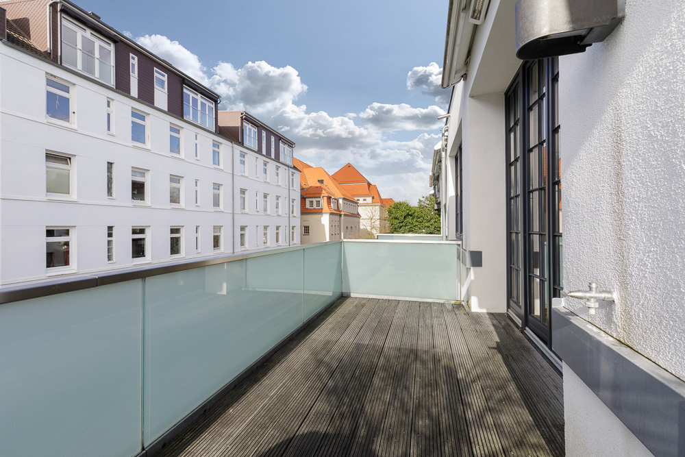 Großer Balkon mit Holzfußboden und Glasgeländer, Blick auf Nachbargebäude und blauen Himmel.
