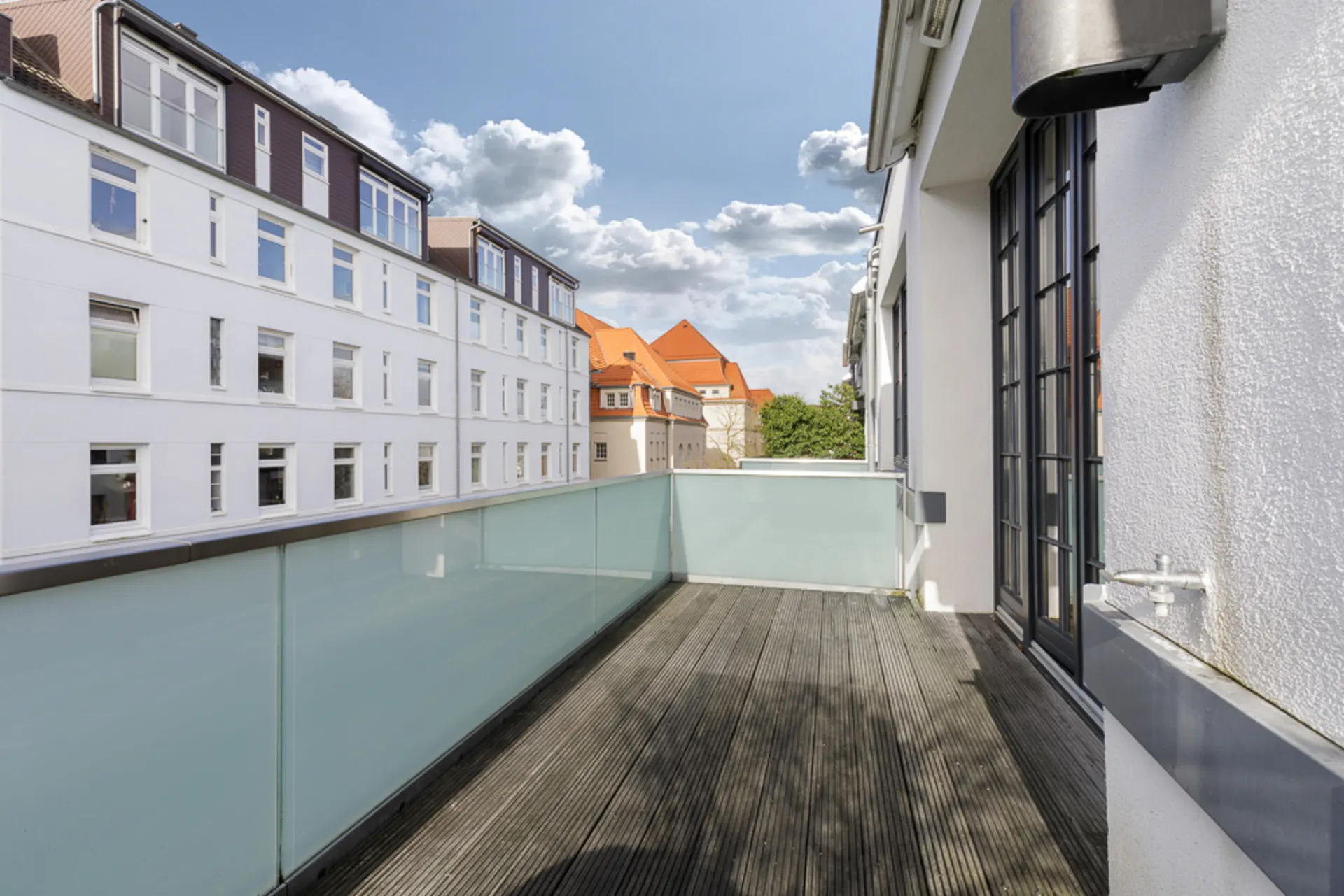 Großer Balkon mit Holzfußboden und Glasgeländer, Blick auf Nachbargebäude und blauen Himmel.