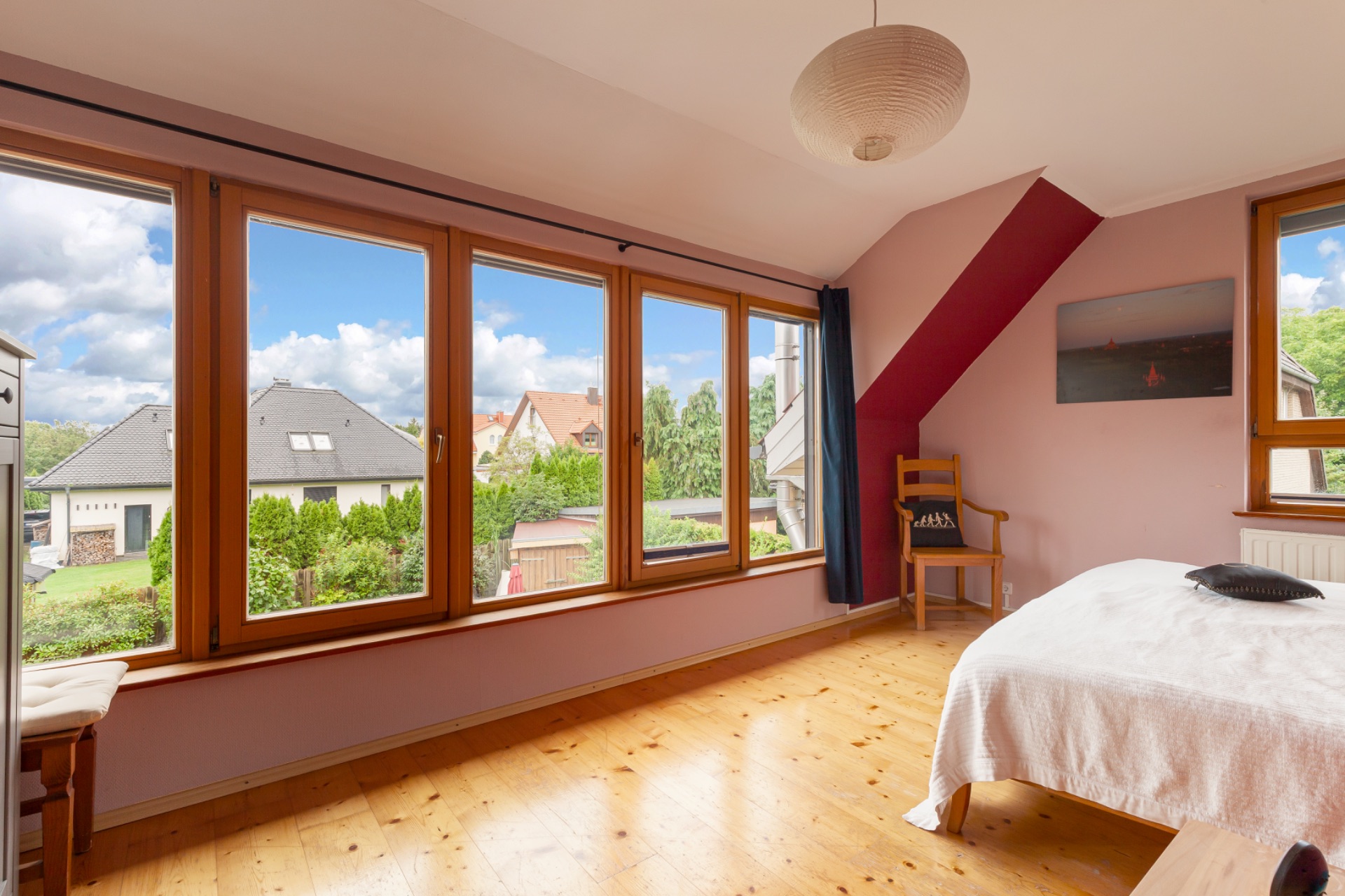 Helles Schlafzimmer mit großen Fenstern, Holzboden, Bett und Stuhl. Blick auf Nachbarhäuser und Garten.