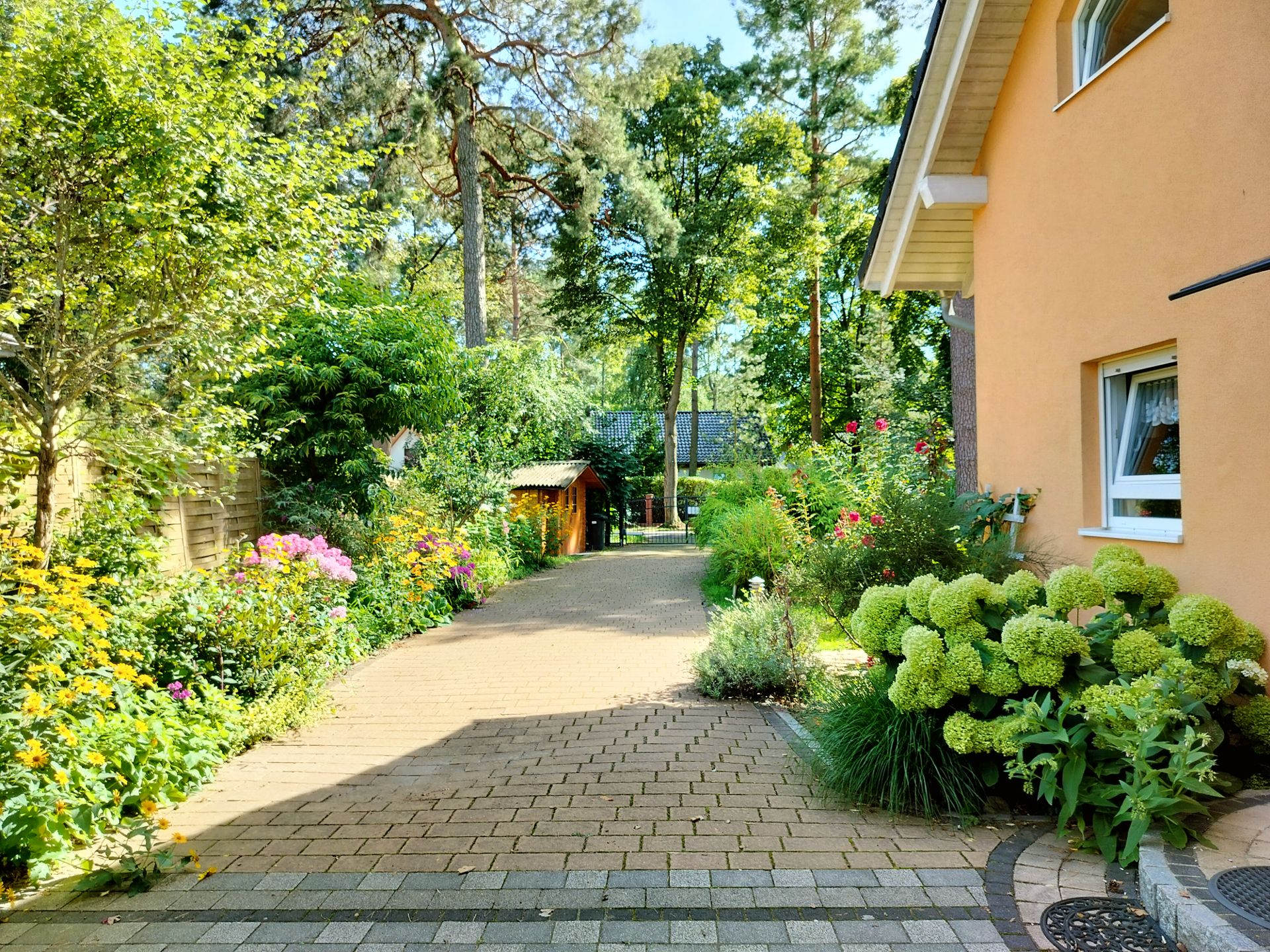 Gepflasterter Weg neben einem gelben Wohnhaus, umgeben von bunten Blumen und Bäumen.