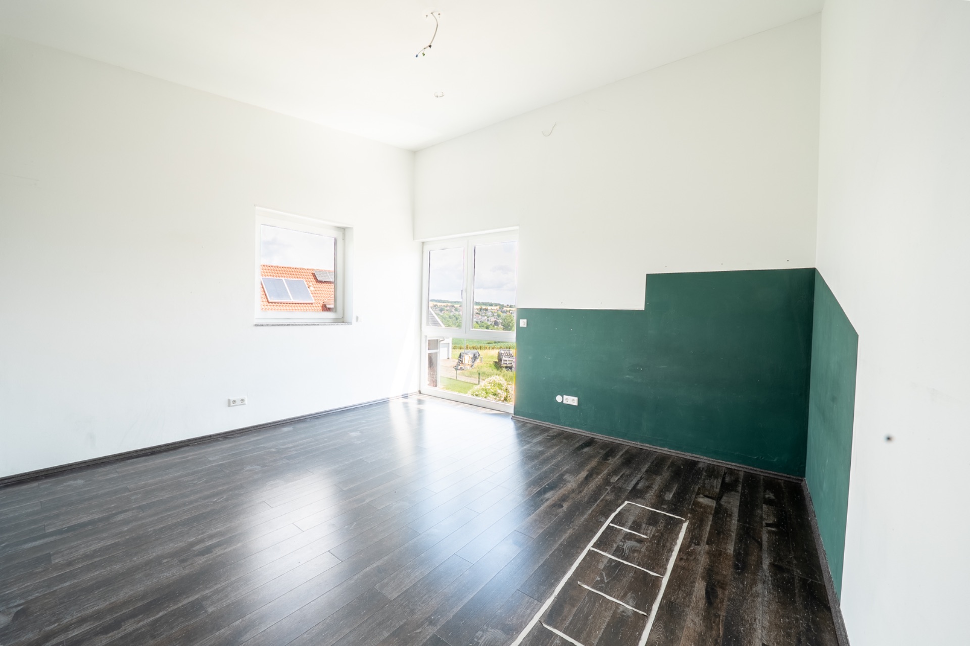 Helles, leeres Zimmer mit dunklem Holzboden, grün gestrichener Wand und Fenster mit Blick ins Freie.