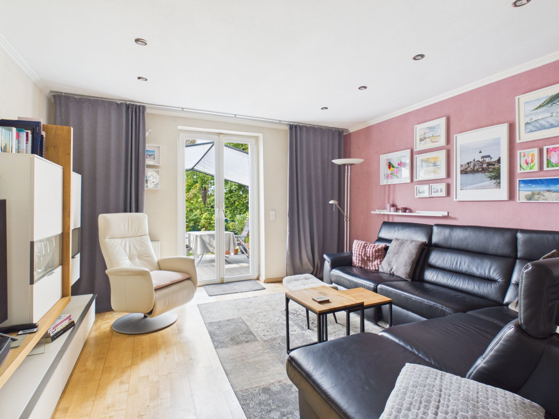 Helles Wohnzimmer mit schwarzem Sofa, Sessel, Couchtisch und Bilderwand, Zugang zur Terrasse.