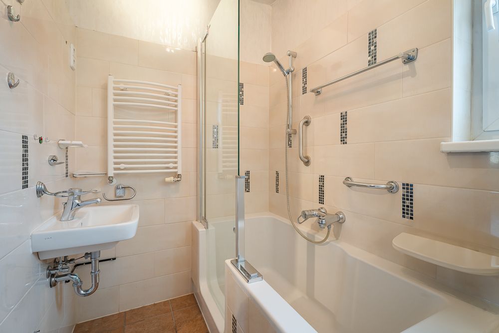 Kleines Badezimmer mit Badewanne, Dusche, Waschbecken und Haltegriffen an den Wänden.