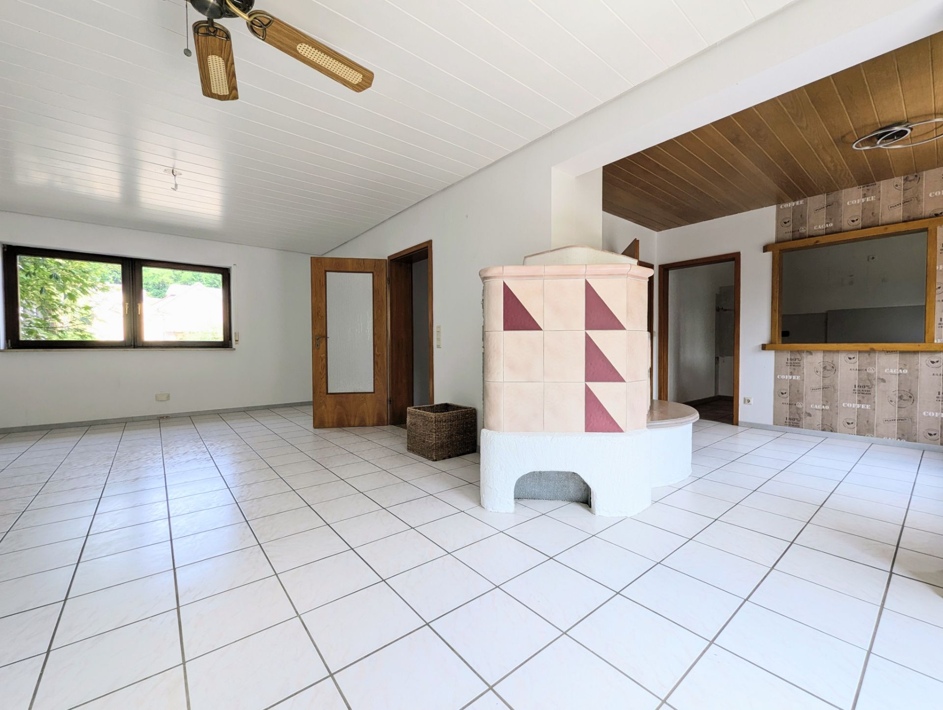 Helles Wohnzimmer mit Kachelofen, Fliesenboden, Holzdecke und großen Fenstern.