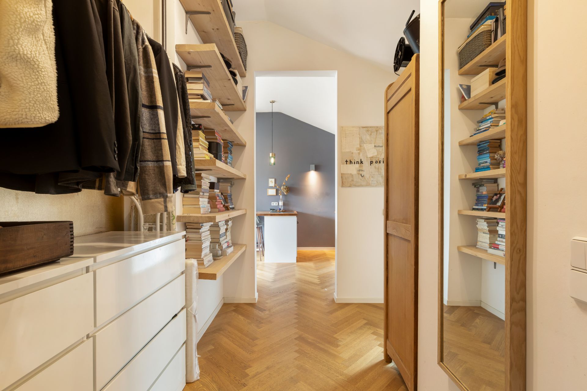 Begehbarer Kleiderschrank mit Regalen, Kleidung, Büchern und Spiegel, Holzboden, Blick in anderen Raum.