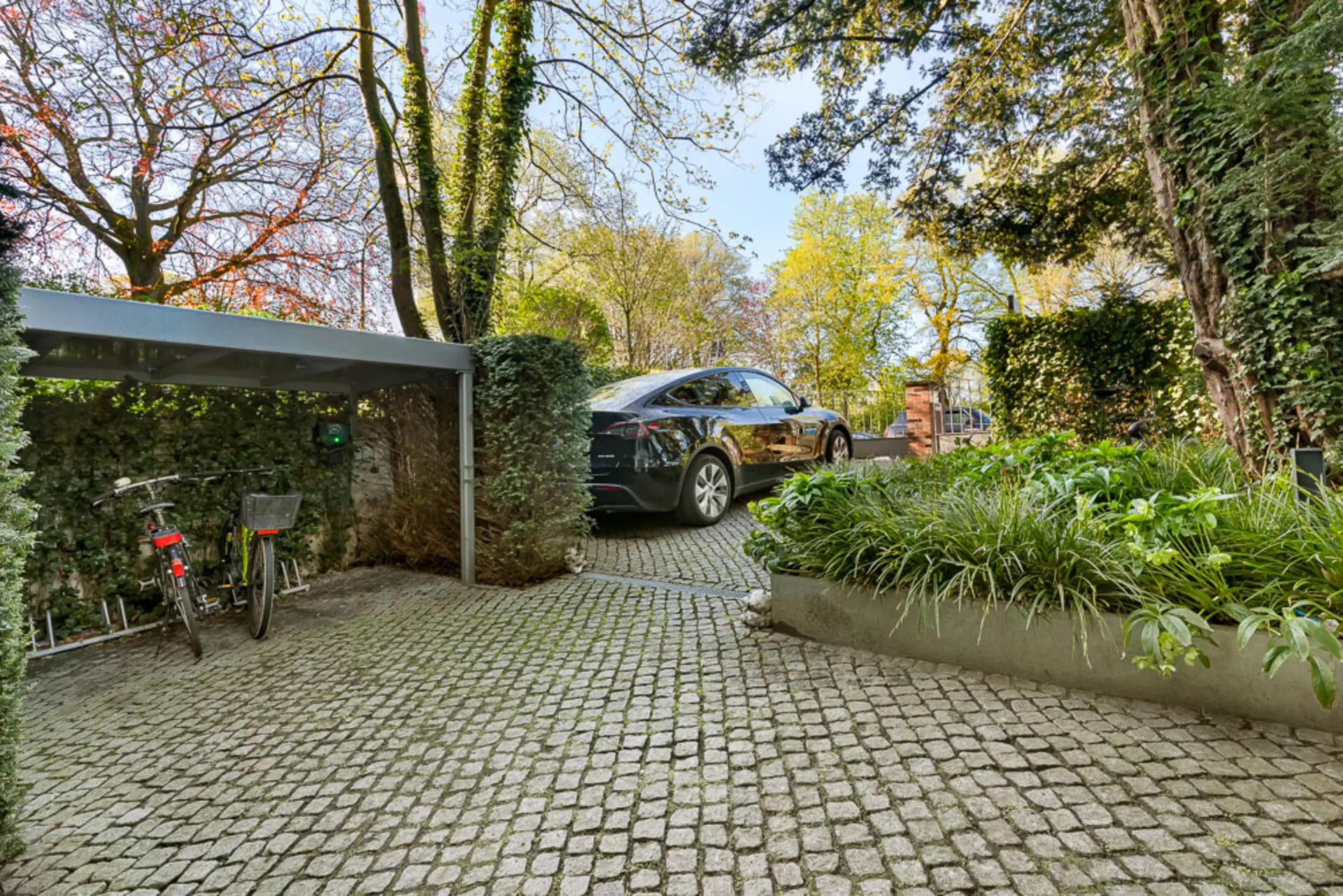 Gepflasterter Hof mit Carport, zwei Fahrrädern, geparktem Auto und viel Grün.