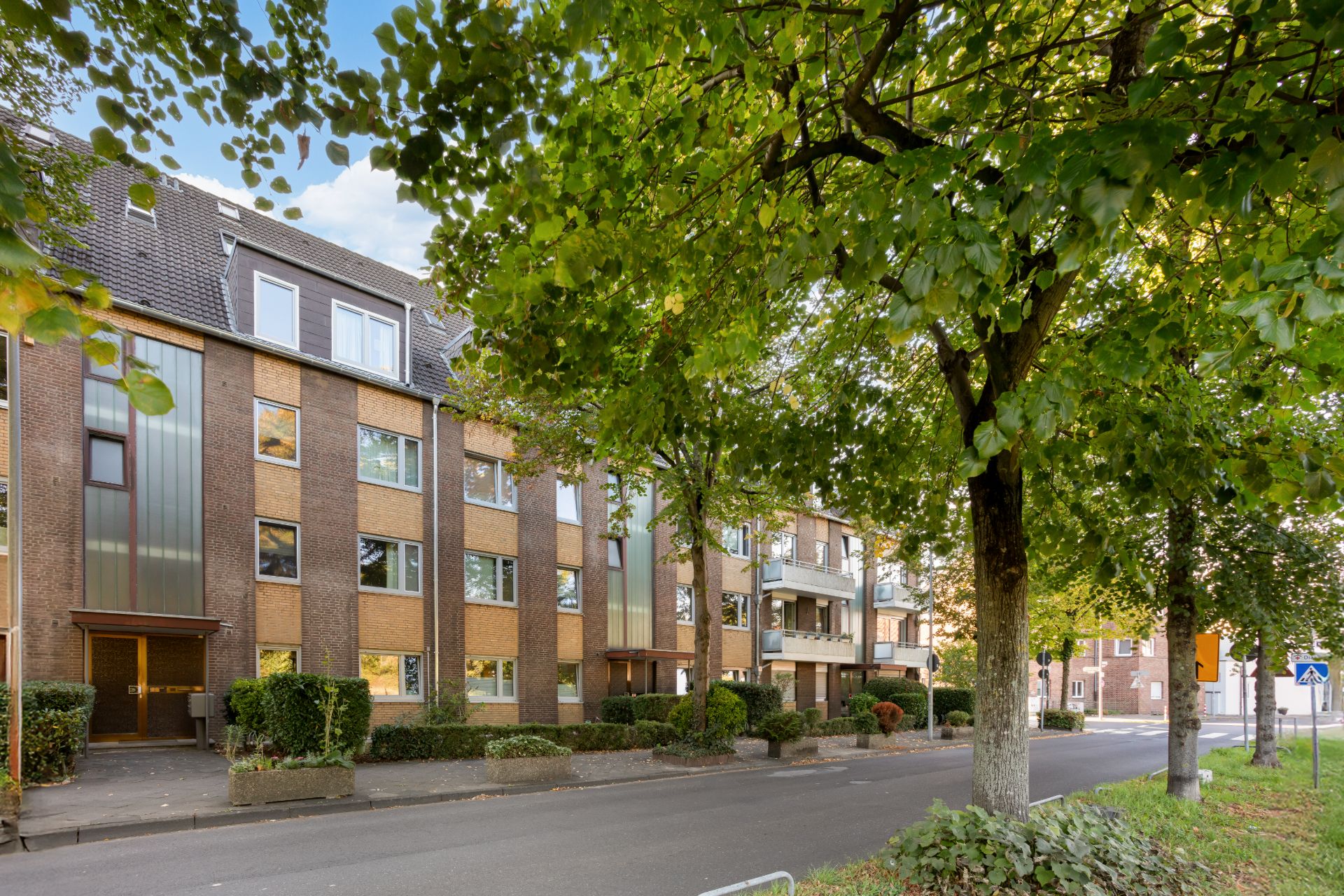 Mehrfamilienhaus aus Backstein an einer von Bäumen gesäumten Straße.