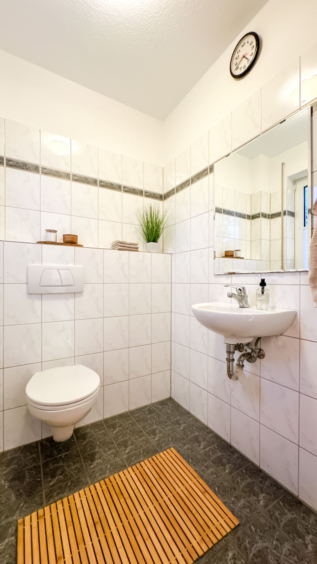 Modernes, gefliestes Badezimmer mit WC, Waschbecken, Spiegel und Holzmatte.