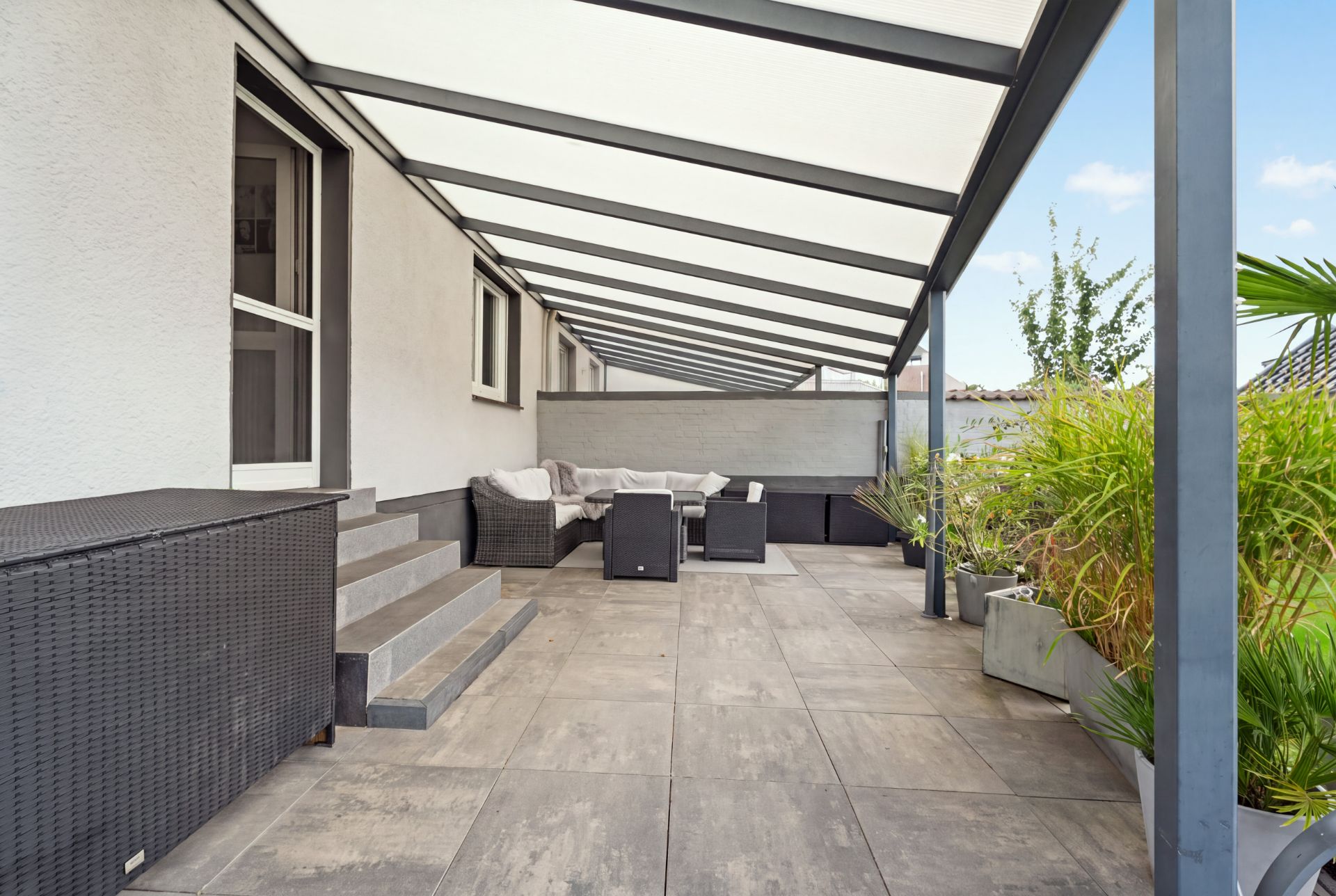 Überdachte Terrasse mit Sitzmöbeln, Pflanzen in Kübeln und Treppe zum Hauseingang.