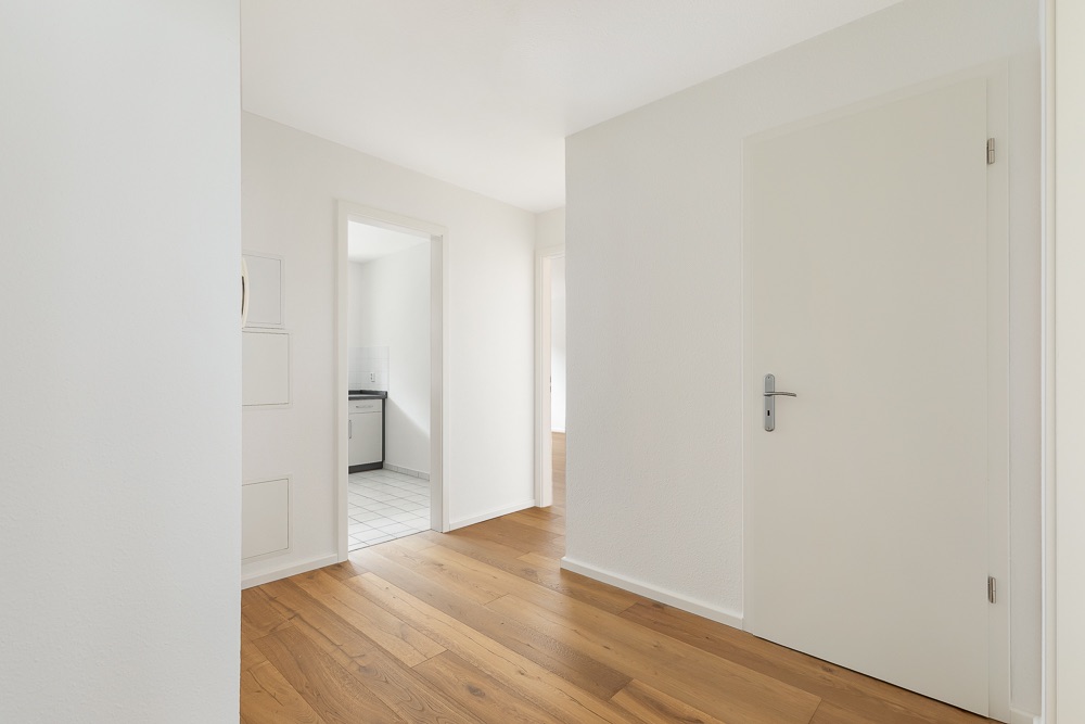 Heller Flur mit Holzboden, weißen Wänden und Blick in Küche mit Fliesenboden.