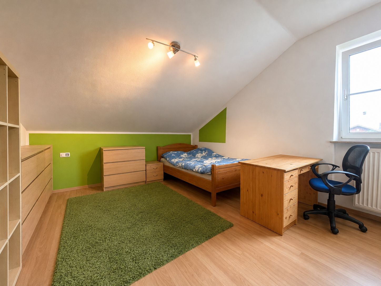 Helles Schlafzimmer mit Einzelbett, Schreibtisch, Stuhl, Kommoden und grünem Teppich auf Holzboden.