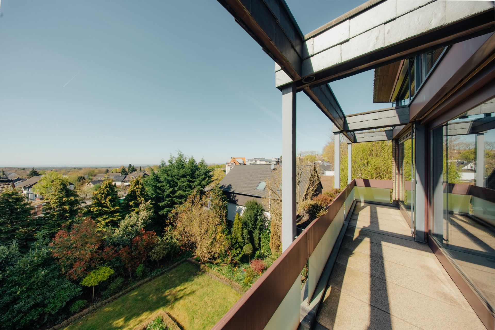 Sonniger Balkon mit Glasgeländer und Blick auf Garten und Nachbarhäuser.