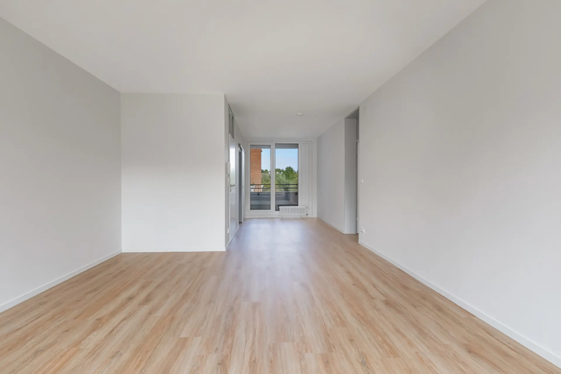 Leerer Raum mit hellem Holzfußboden und weißen Wänden, Fenster am Ende des Zimmers.