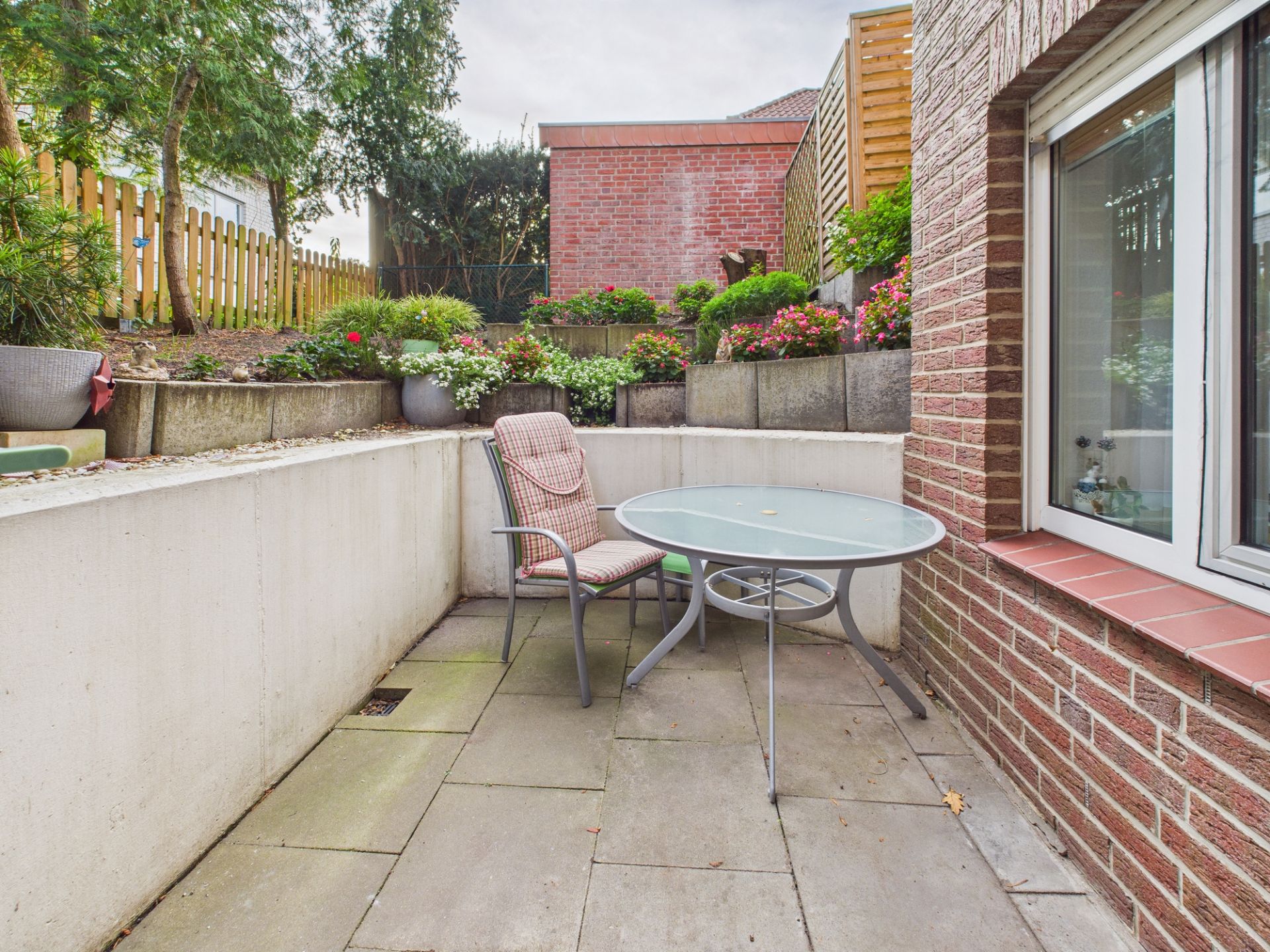 Kleine Terrasse mit Glas­tisch, Stuhl und Blumenbeeten an einer Hauswand.