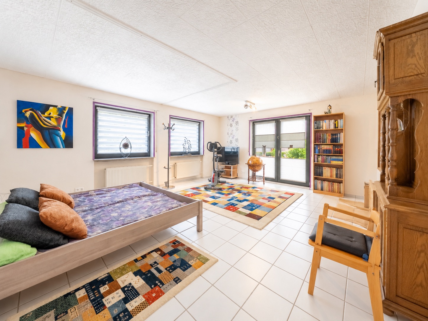Helles Schlafzimmer mit Bett, Holzschrank, Teppichen, Bücherregal und Fenstern.