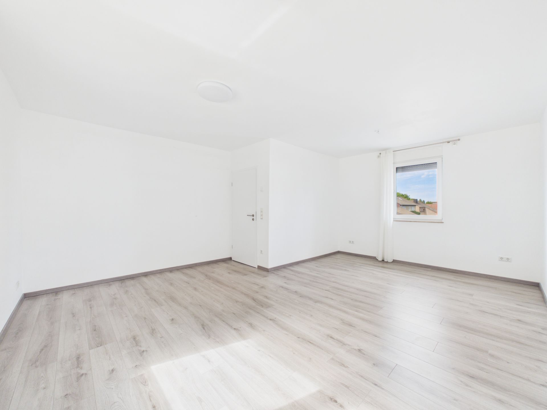 Heller, leerer Raum mit weiß gestrichenen Wänden, Laminatboden und einem Fenster mit Vorhang.