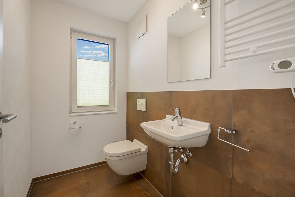 Modernes Badezimmer mit Wand-WC, Waschbecken, Spiegel und Fenster mit Plissee.