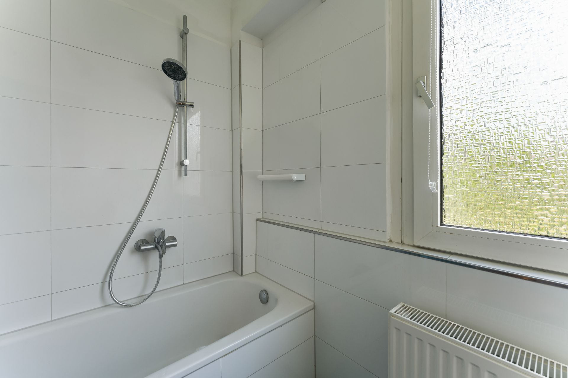 Weiße Badewanne mit Duschkopf, Fliesenwand und Fenster mit Milchglas.