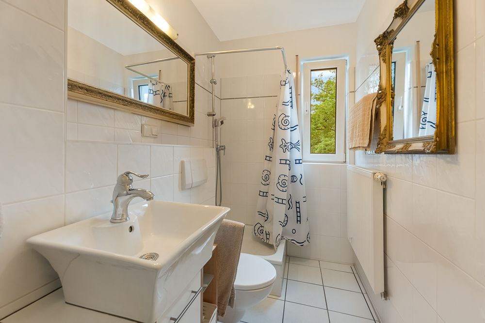 Helles Badezimmer mit Waschbecken, Spiegel, Toilette, Dusche mit Duschvorhang und zwei Fenstern.
