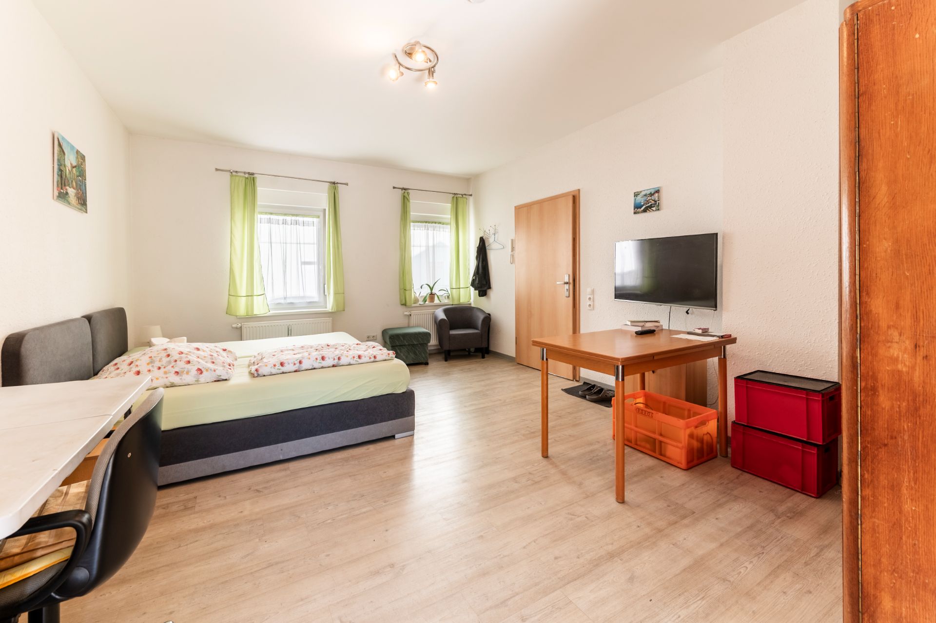 Helles Schlafzimmer mit Doppelbett, Tisch, Fernseher und zwei Fenstern mit grünen Vorhängen.