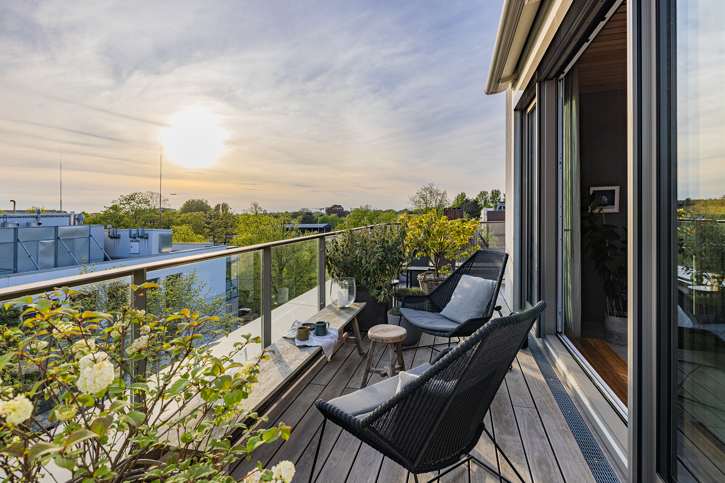 Moderne Balkonterrasse mit Glasgeländer, Pflanzen, Sitzmöbeln und Ausblick auf Bäume bei Sonnenuntergang.