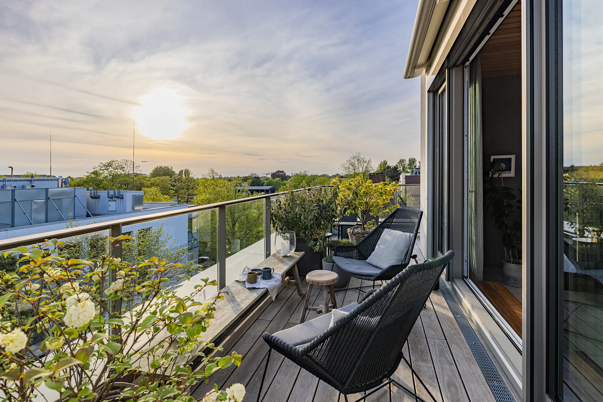 Moderne Balkonterrasse mit Glasgeländer, Pflanzen, Sitzmöbeln und Ausblick auf Bäume bei Sonnenuntergang.