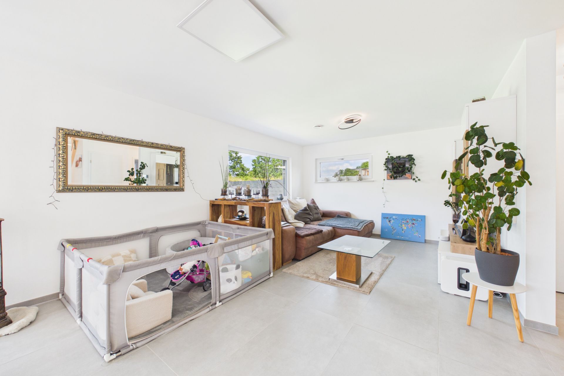 Helles Wohnzimmer mit Ecksofa, Spielgehege und großen Fenstern, viele Pflanzen und moderner Dekor.