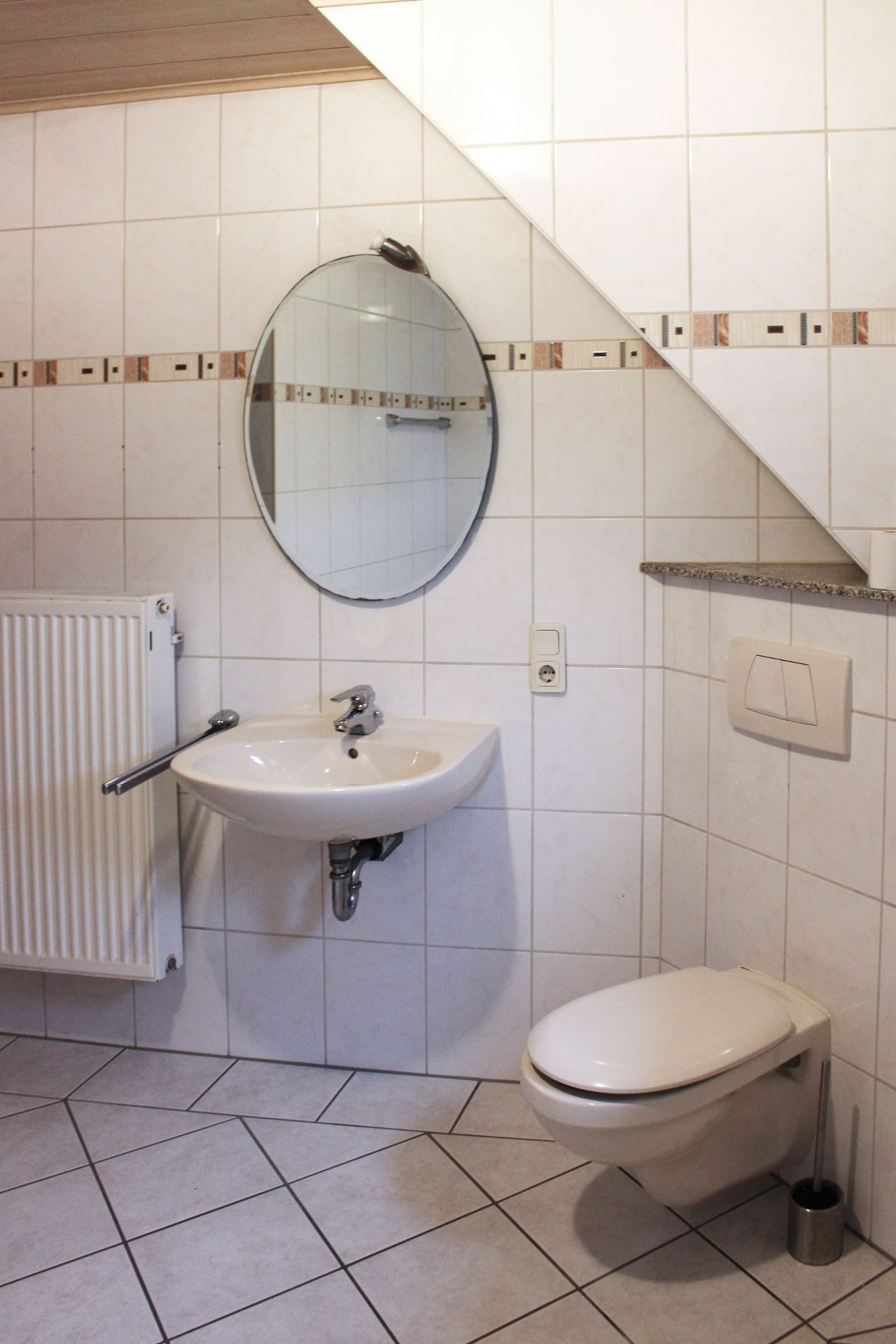 Gefliestes Badezimmer mit Waschbecken, rundem Spiegel und Wandtoilette unter Dachschräge.