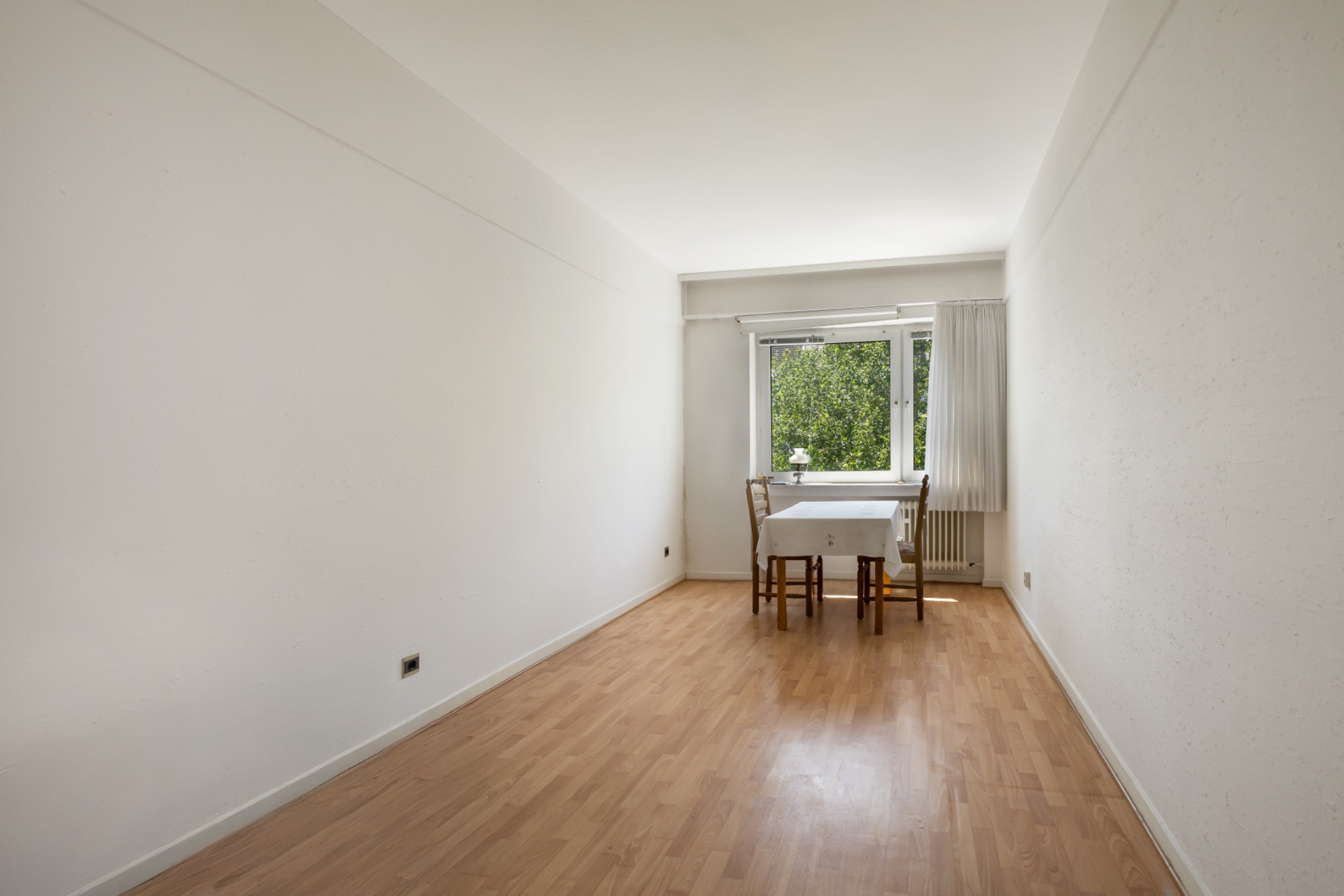 Helles Zimmer mit Holzfußboden, Tisch und zwei Stühlen vor einem Fenster mit Blick ins Grüne.