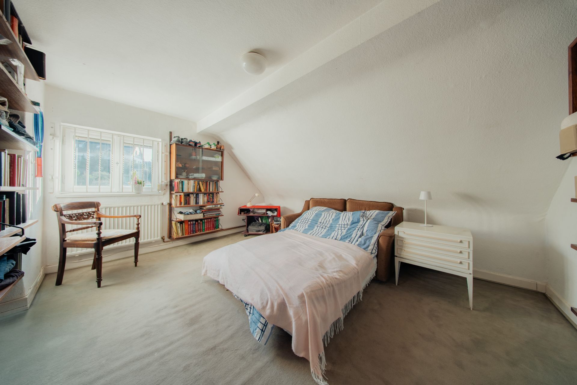 Helles Schlafzimmer mit Bett, Bücherregalen, Sessel und Fenster unter einer Dachschräge.