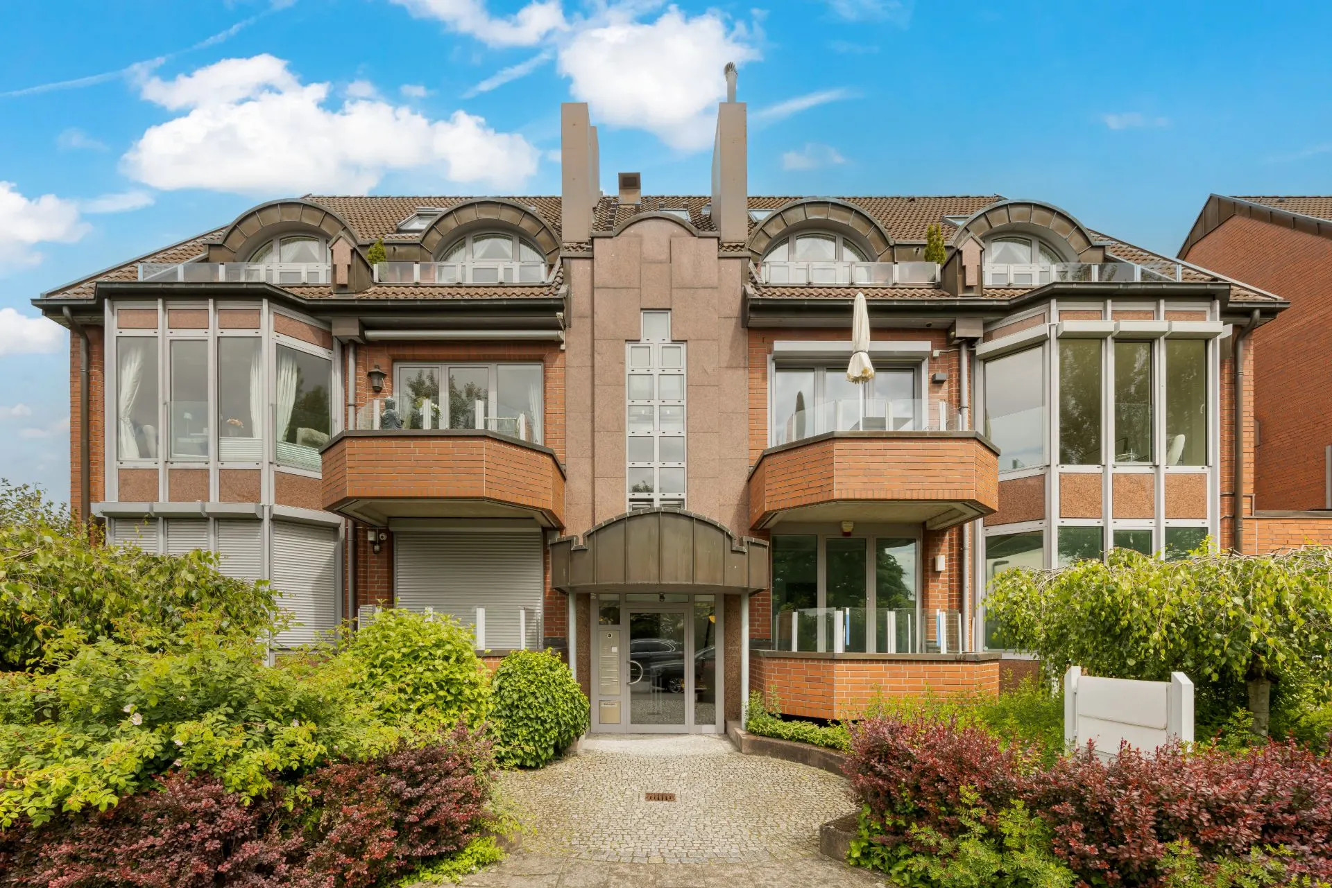 Moderne Wohnhausfassade mit großen Fenstern, Balkonen und bepflanztem Vorgarten.